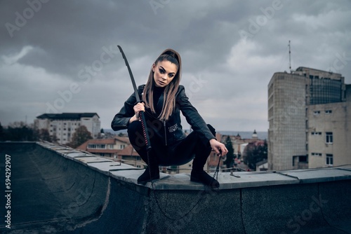 Cool female ninja warrior in leather outfit wielding a Japanese sword on the top of a building photo