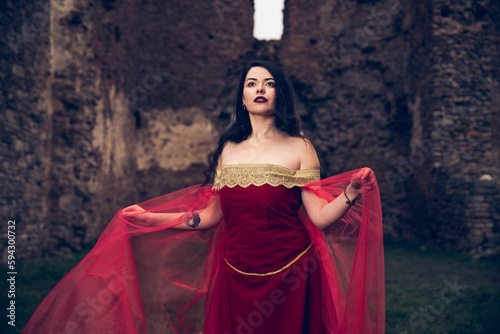 Young beautiful female in medieval red dress posing near old ruins