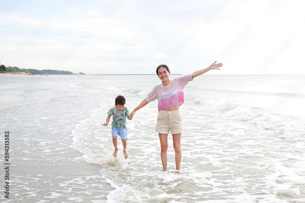 Happy Asian mom and son at tropical sand beach with waving hands. Happy family mother and little boy having fun in summer holiday.