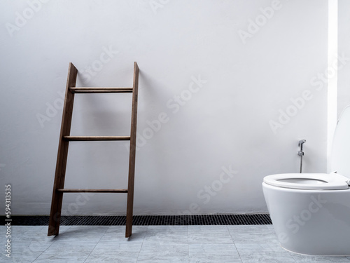 White restroom interior design, minimal style. Wooden towel rack, dark brown standing towel ladder near ceramic flush toilet and bidet shower on square tiles and white wall background with copy space.