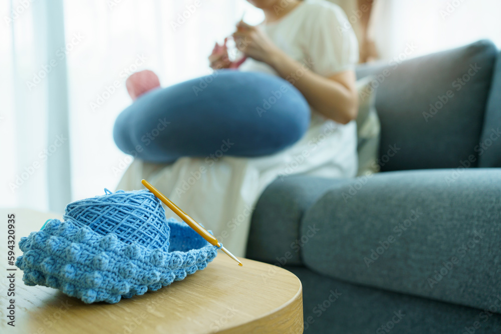 Fototapeta premium Happy cheerful Asian woman doing a crochet in living room in free time. Skillful Asian craftswoman knitting a handcraft crochet sweater and hat in cozy living room.