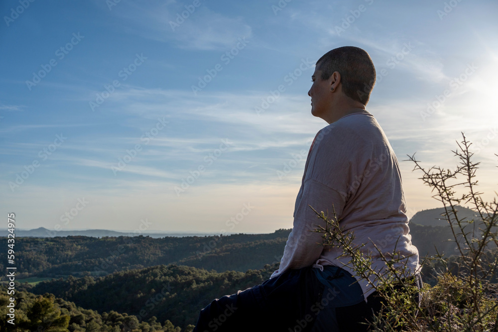 Chubby non-binary woman in profile on a cliff with a beautiful view of nature at sunset, pensive and looking alone at the landscape.
