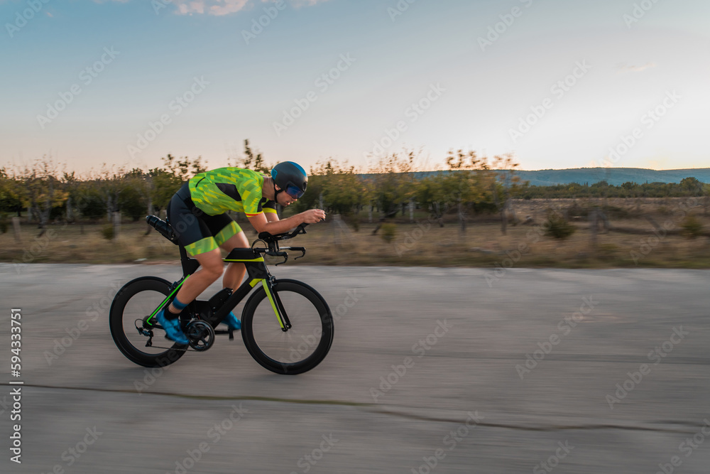 Obraz premium Triathlete riding his bicycle during sunset, preparing for a marathon. The warm colors of the sky provide a beautiful backdrop for his determined and focused effort.