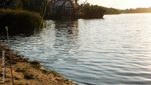 River and ships at sunset. Calm waves of the lake wash the sandy shore and reeds. Ship with industrial equipment works on a pond. Snails and seaweed on the seashore. Beautiful water landscape. Nature photo