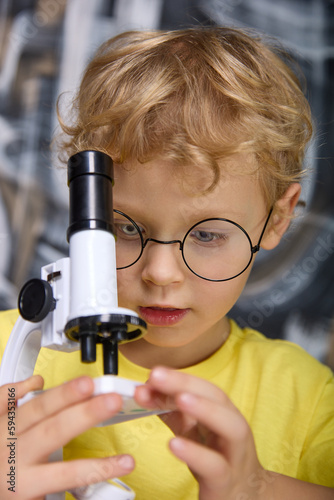 Close-up of a cute little kid wearing glasses and a yellow T-shirt emitting a substance using a microscope. In the hands of a boy with glasses, a real microscope with a black tube