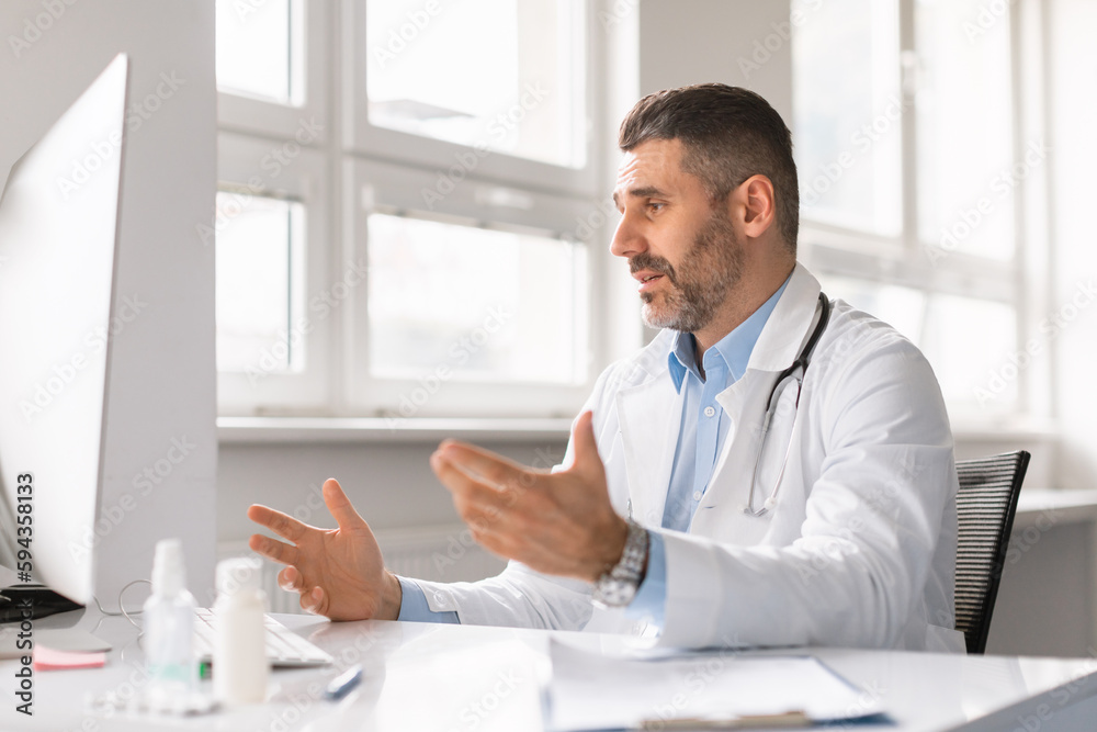 Confident man doctor working on computer at clinic, looking at screen, gesturing, having video conference with patient Confident man doctor working on computer at clinic, looking at screen, gesturing, having video conference with patient