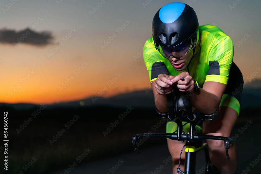 Naklejka premium A triathlete rides his bike in the darkness of night, pushing himself to prepare for a marathon. The contrast between the darkness and the light of his bike creates a sense of drama and highlights the