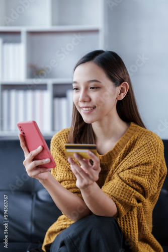 Using mobile cellphone  Young attractive asian female housewife sitting on sofa using credit card  buying online shopping purchase stay at home
