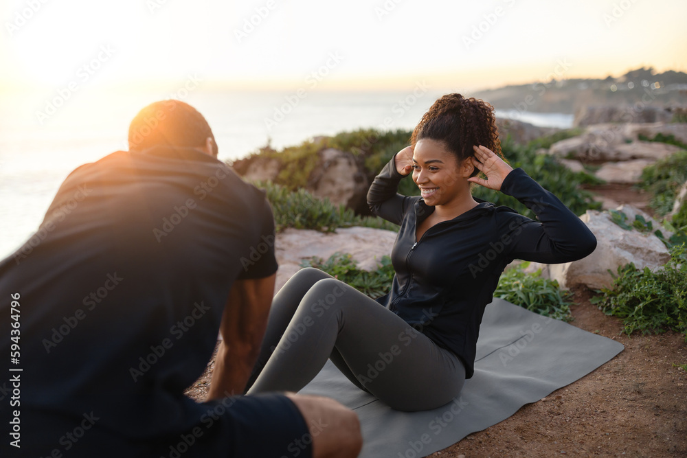 Smiling millennial african american man help to lady in sportswear do abdominal exercises on ocean beach