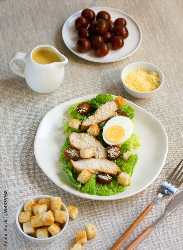 Caesar salad with croutons, fried chicken, cherry tomatoes, egg and parmesan cheese on a light plate with a fork, on a linen tablecloth in a plate of grated parmesan cheese and cherry tomatoes
