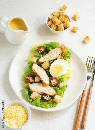 Caesar salad with croutons, fried chicken, cherry tomatoes, egg and parmesan cheese on a light plate, on a light table against the background of a bowl with croutons, grated parmesan cheese