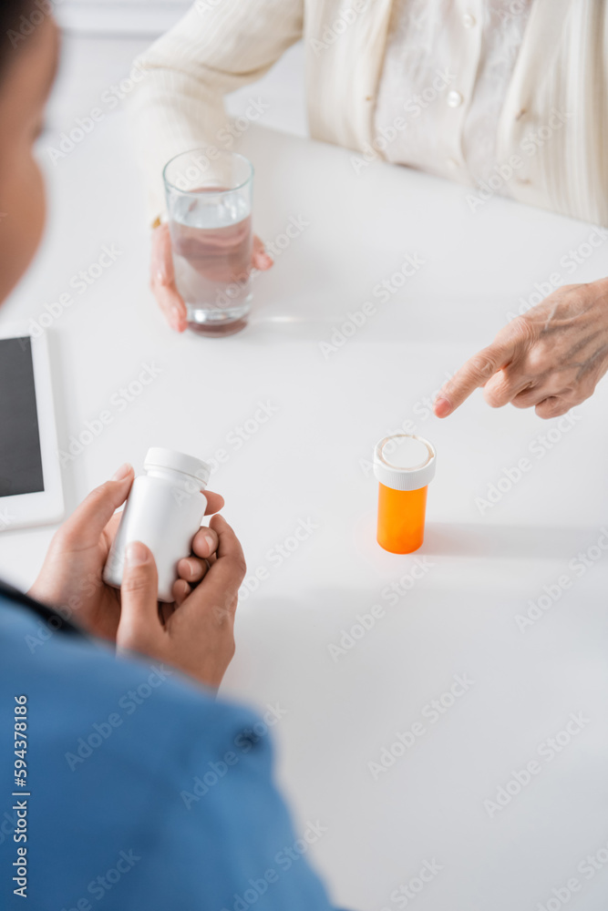 cropped view of retired woman pointing at bottle with medication near multiracial nurse on blurred foreground. cropped view of retired woman pointing at bottle with medication near multiracial nurse on blurred foreground.
