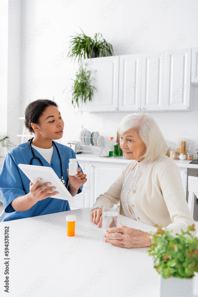Fototapeta premium brunette multiracial nurse using digital tablet and holding medication while talking to retired woman with grey hair.