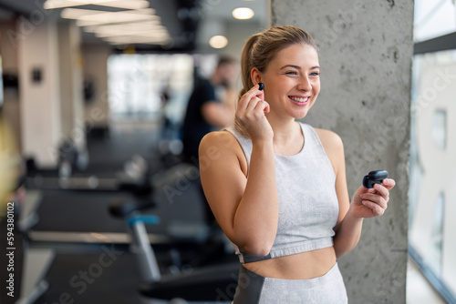 Young beautiful sportswoman with earphones. Smiling fitness woman putting on in-ear headphones  holding charging case. Happy female athlete listening to music over earphones  enjoying her time in gym.