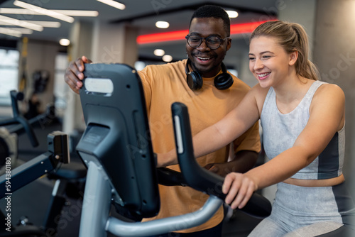 Confident male personal trainer cheerful athlete adjusting training mode on exercise gym to fit young woman smiling gym client, assisting her, giving advice for effective cardio training in gym.