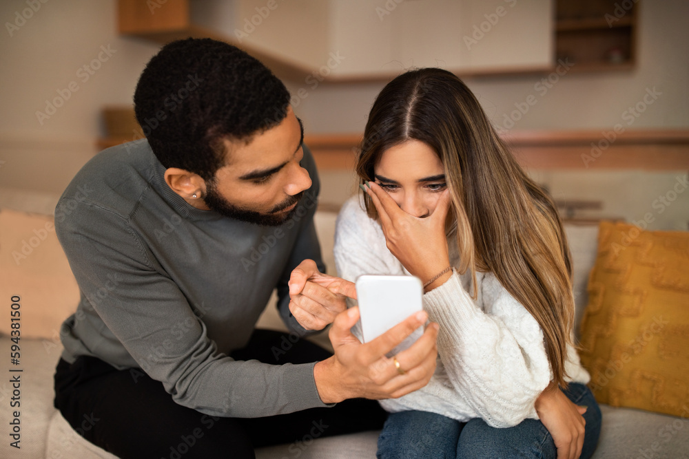 Angry millennial middle eastern husband showing smartphone to frightened caucasian wife quarreling Angry millennial middle eastern husband showing smartphone to frightened caucasian wife quarreling