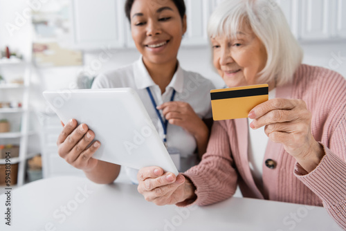 brunette multiracial social worker holding digital tablet near happy senior woman with credit card.