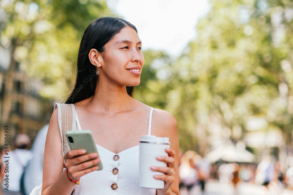 Glad Latin American woman with cellphone and zero waste cup of coffee to go smiling and looking away on blurred background of sunlit park in Barcelona, Spain Glad Latin American woman with cellphone and zero waste cup of coffee to go smiling and looking away on blurred background of sunlit park in Barcelona, Spain