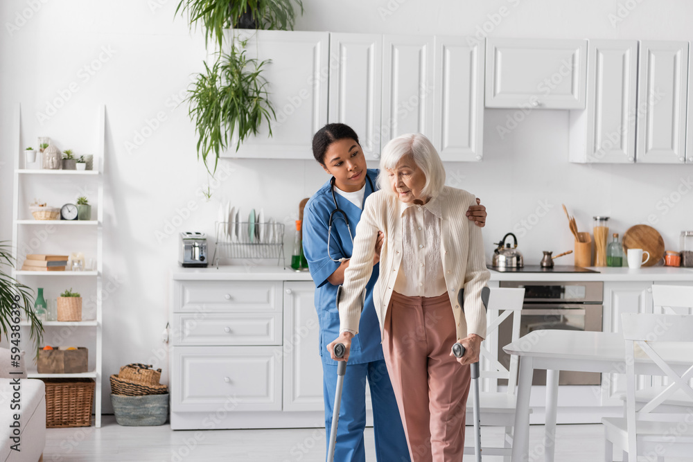 © LIGHTFIELD STUDIOS - brunette multiracial caregiver in blue uniform helping retired woman using crutches to walk in apartment. © LIGHTFIELD STUDIOS - brunette multiracial caregiver in blue uniform helping retired woman using crutches to walk in apartment.