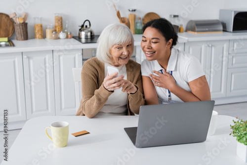 cheerful multiracial social worker looking at smartphone in hands of retired woman near laptop.