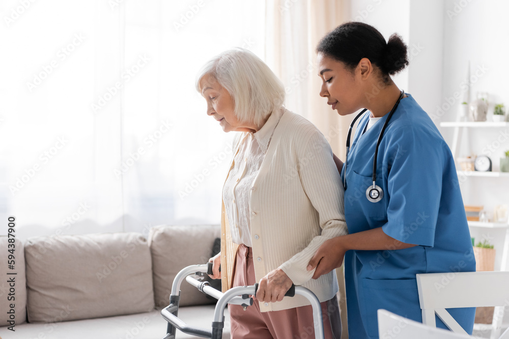 Fototapeta premium brunette multiracial nurse in uniform supporting retired woman with grey hair walking with walker at home.