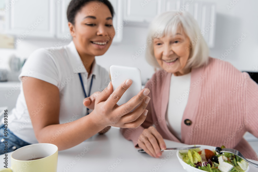 happy multiracial social worker showing smartphone to senior woman during lunch in kitchen.