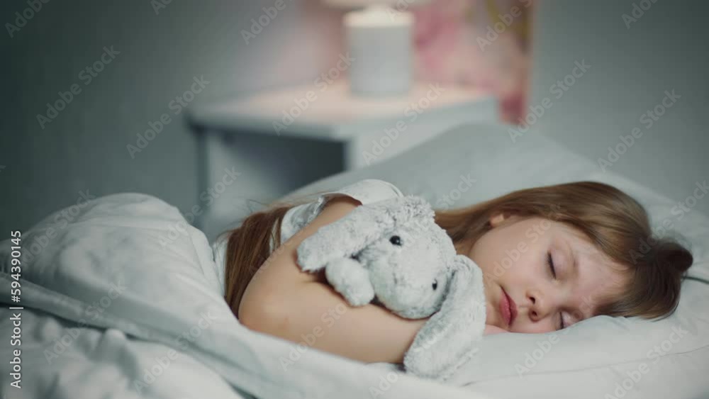 Cute funny cheerful little girl with brown hair sleeping in the white bed with favorite fluffy toy bunny. 