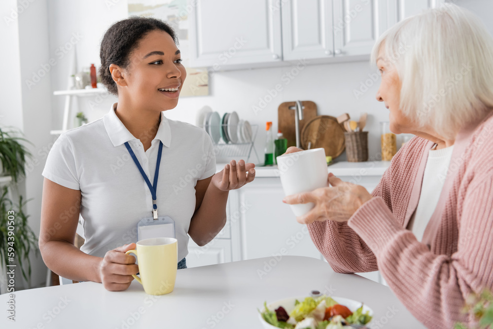 happy multiracial social worker having conversation with senior woman over cups of tea. happy multiracial social worker having conversation with senior woman over cups of tea.