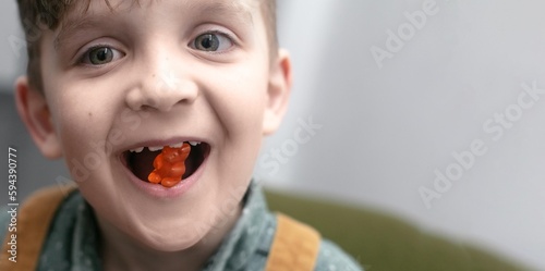 Close-up portrait of a cute boy 7 years old, holding a gummy bear in his mouth. Children's vitamins to improve immunity.