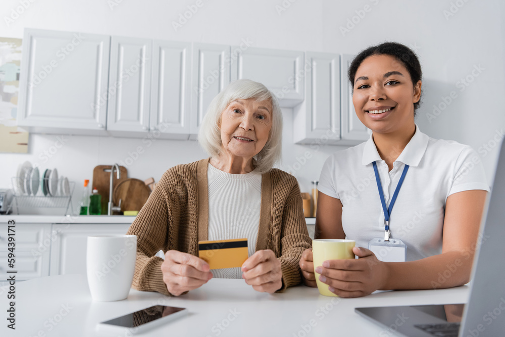 cheerful multiracial social worker holding cup near senior woman with credit card and gadgets. cheerful multiracial social worker holding cup near senior woman with credit card and gadgets.