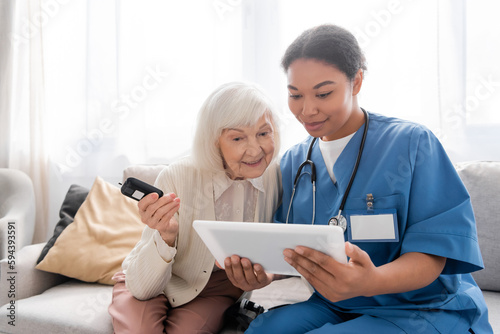 cheerful senior woman with grey hair holding glucometer and looking at digital tablet near happy multiracial nurse.