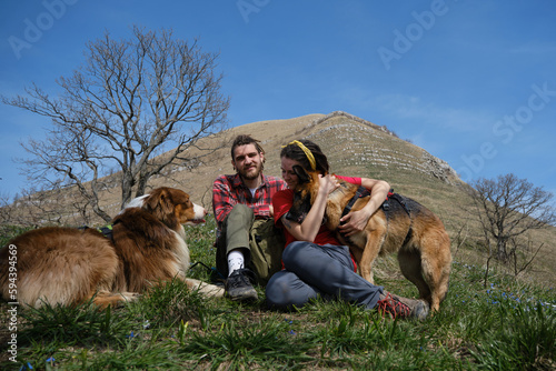 Couple of travelers sitting at foot of mountain in clearing among spring flowers with two dogs. Australian and German Shepherds travel with man and woman pet owners. Concept hiking with dog.