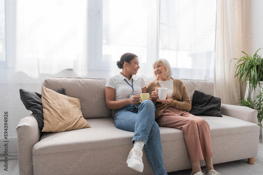 happy multiracial social worker having tea with senior woman in living room.