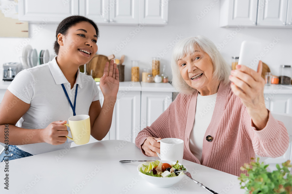 happy senior woman holding smartphone while cheerful multiracial social worker waving hand during video call. happy senior woman holding smartphone while cheerful multiracial social worker waving hand during video call.