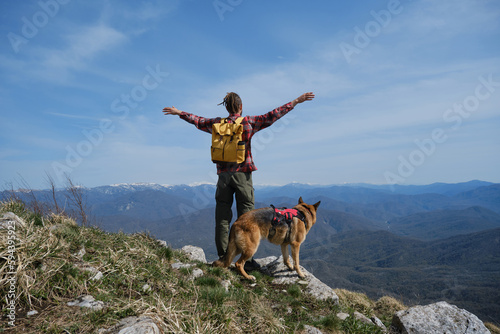 Traveler with yellow backpack standing on top of mountain and raised hands up in different directions, German shepherd standing next to him. Nature of Caucasus. Rear view. Concept hiking with dog.