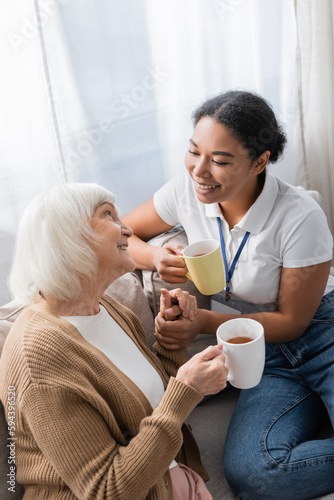 cheerful multiracial social worker chatting with senior woman while having tea in living room. photo