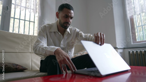 Young man sitting on couch and opening laptopscreen at home. One Arab Middle Eastern person browsing internet online with modern technology photo