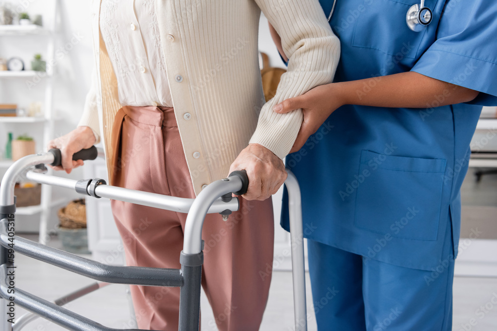 cropped view of multiracial nurse in blue uniform supporting senior woman walking with help of walker at home.