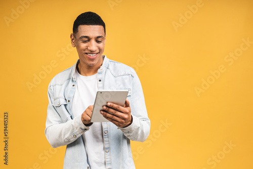 Working on digital tablet. Portrait of happy smiling young African american man holding digital tablet while standing isolated over yellow background.