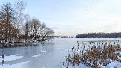In winter, in clear and frosty weather, smooth ice forms on the lake. Reeds are frozen into the ice. Snow lies on the banks and in places on the ice. There is a road with a bridge across the channel