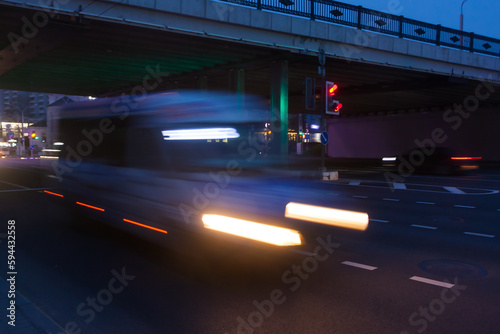 A blurred minibus moves under an overpass through the city in the evening
