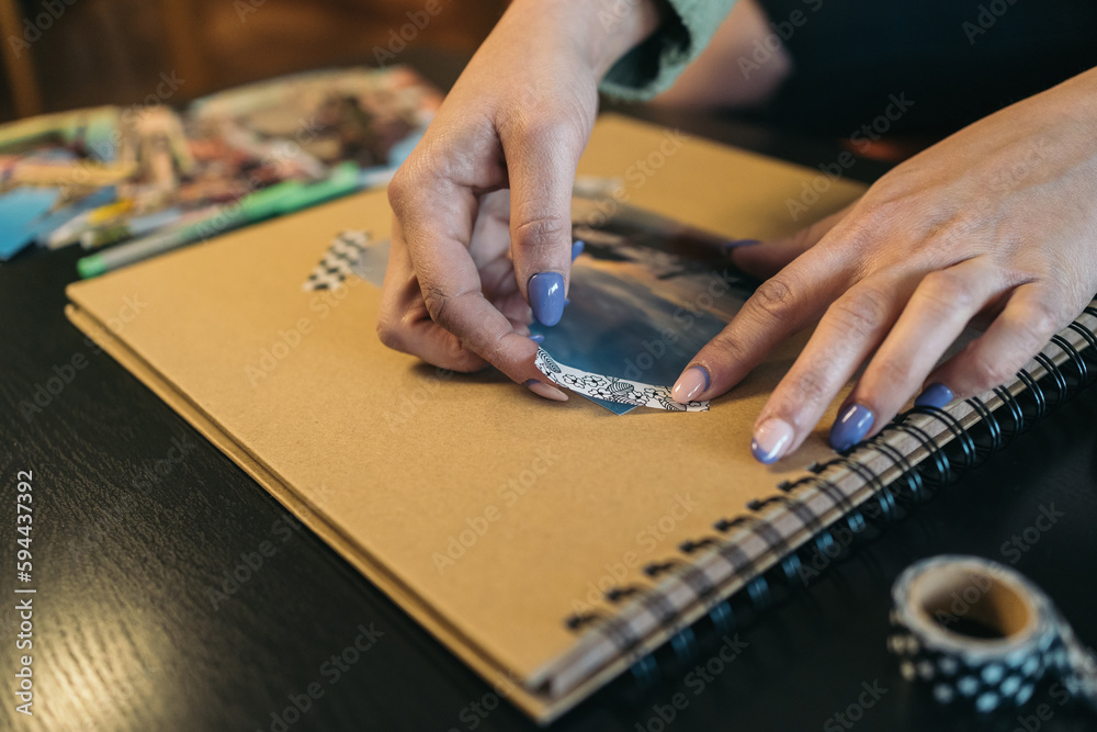 Fototapeta premium Middle-aged woman's hands sticking a small piece of washi tape on the cover of a handmade travel kraft paper photo album.