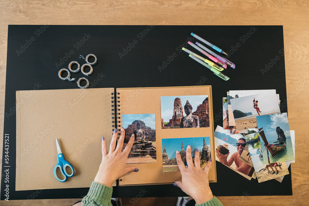 Image seen from above of a middle-aged woman's hands placing the photos and markers to make her handmade travel kraft paper photo album. - obrazy, fototapety, plakaty 