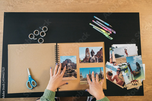 Image seen from above of a middle-aged woman's hands placing the photos and markers to make her handmade travel kraft paper photo album. photo