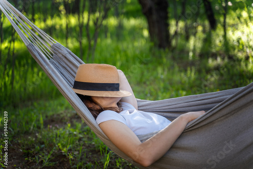 Woman resting in a hammock in a summer garden covering her face with a straw hat. Summer relax vacation