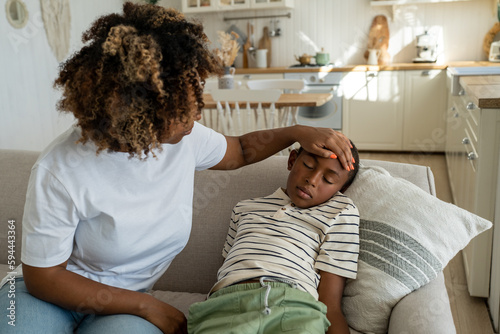 Fever in kids. African American mother touching forehead of sick little boy son at home, caring parent mom checking childs temperature with hand. Schoolboy lying on sofa feeling unwell and ill