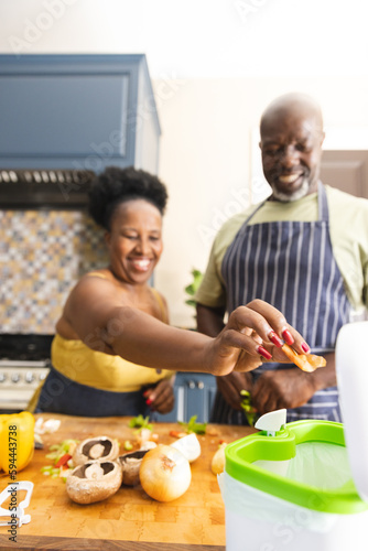 Happy senior african american couple wearing aprons and throwing waste out in kitchen