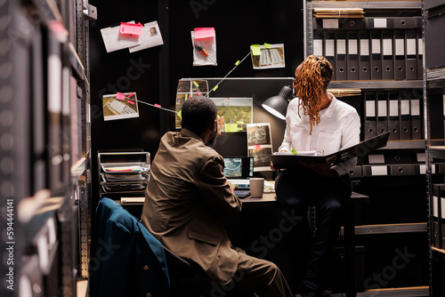 Detective partners analyzing surveillance photos hanging on evidence board and working late. African american man and woman cops studying crime scene photographs and forensic reports