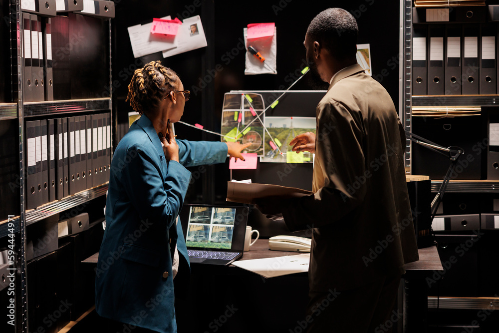 Police detective partners analyzing clues scheme on detective board. African american woman cop pointing at crime scene and suspect photos connection map, discussing insight with investigator Police detective partners analyzing clues scheme on detective board. African american woman cop pointing at crime scene and suspect photos connection map, discussing insight with investigator