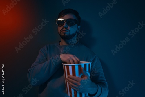 A man is in a dark room with neon lighting, watching a movie with 3D glasses and popcorn.